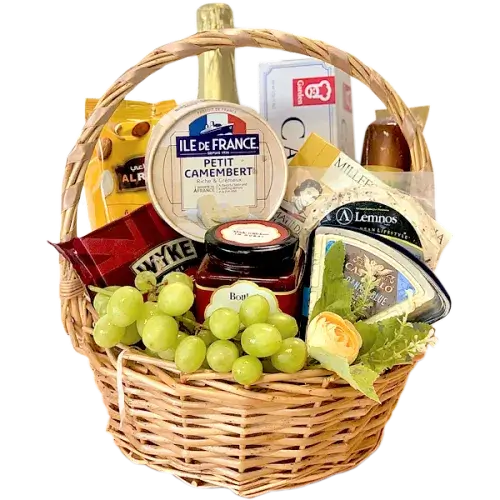 A photo of a wicker gift basket overflowing with a variety of cheeses, grapes, nuts, crackers, a jar of chutney, a bottle of sparkling grape juice, and small hazelnut snacks.