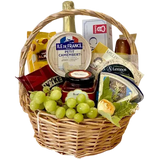 A photo of a wicker gift basket overflowing with a variety of cheeses, grapes, nuts, crackers, a jar of chutney, a bottle of sparkling grape juice, and small hazelnut snacks.