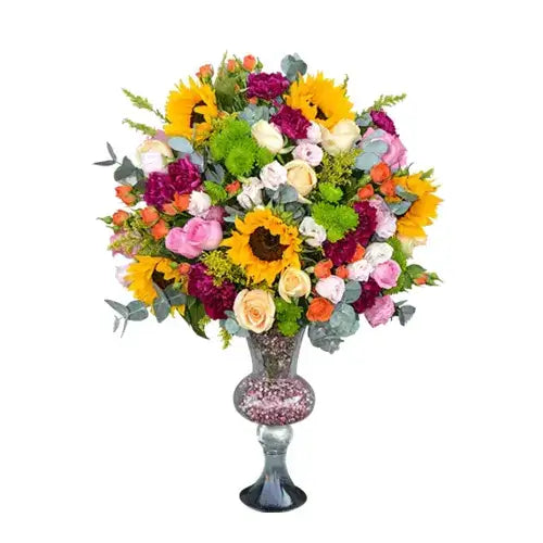 Mixed flower bouquet with sunflowers, peach roses, pink roses, purple carnations, pink lisianthus, spray orange roses, and chrysanthemums in a vase.