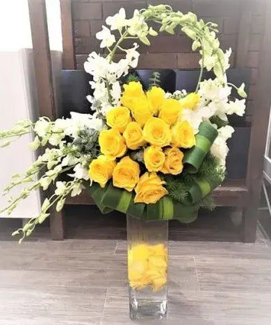 Tall flower arrangement with yellow roses, white orchids, eucalyptus, and greenery in a clear glass vase decorated with yellow rose petals