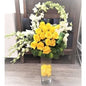 Tall flower arrangement with yellow roses, white orchids, eucalyptus, and greenery in a clear glass vase decorated with yellow rose petals