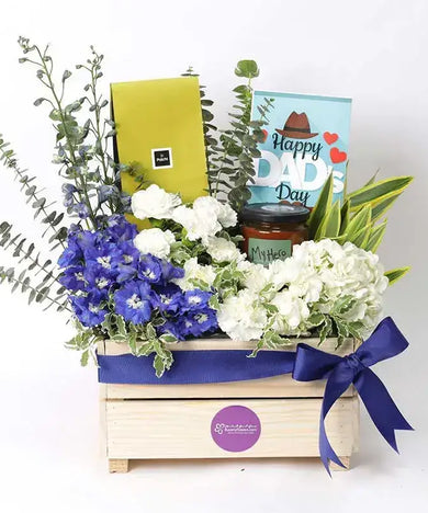 a Father's Day gift hamper with a wooden box overflowing with blue and white flowers, Patchi chocolates, a greeting card, and a scented candle
